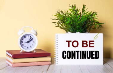 Renkli günlüklerin üzerinde duran yeşil bir bitki ve beyaz alarmlı bir saatin yanındaki bir deftere yazılmıştır..