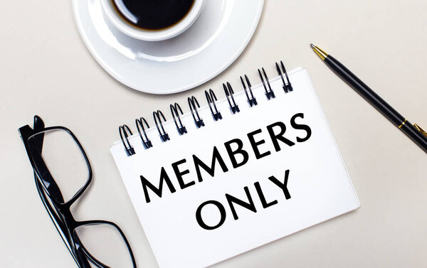 Очки, белая чашка кофе, белая тетрадь со словами "ТОЛЬКО МЕМБЕРЫ" и шариковая ручка лежат на светлом фоне. Плоский лежал. Вид сверху.