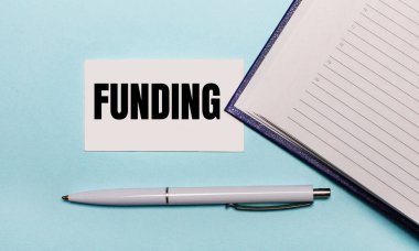 Açık mavi bir arkaplanda, açık bir defter, beyaz bir kalem ve üzerinde yazı yazan bir kart. Yukarıdan görüntüle