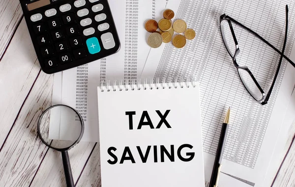 TAX SAVING written in a white notepad near a calculator, cash, glasses, a magnifying glass and a pen. Business concept