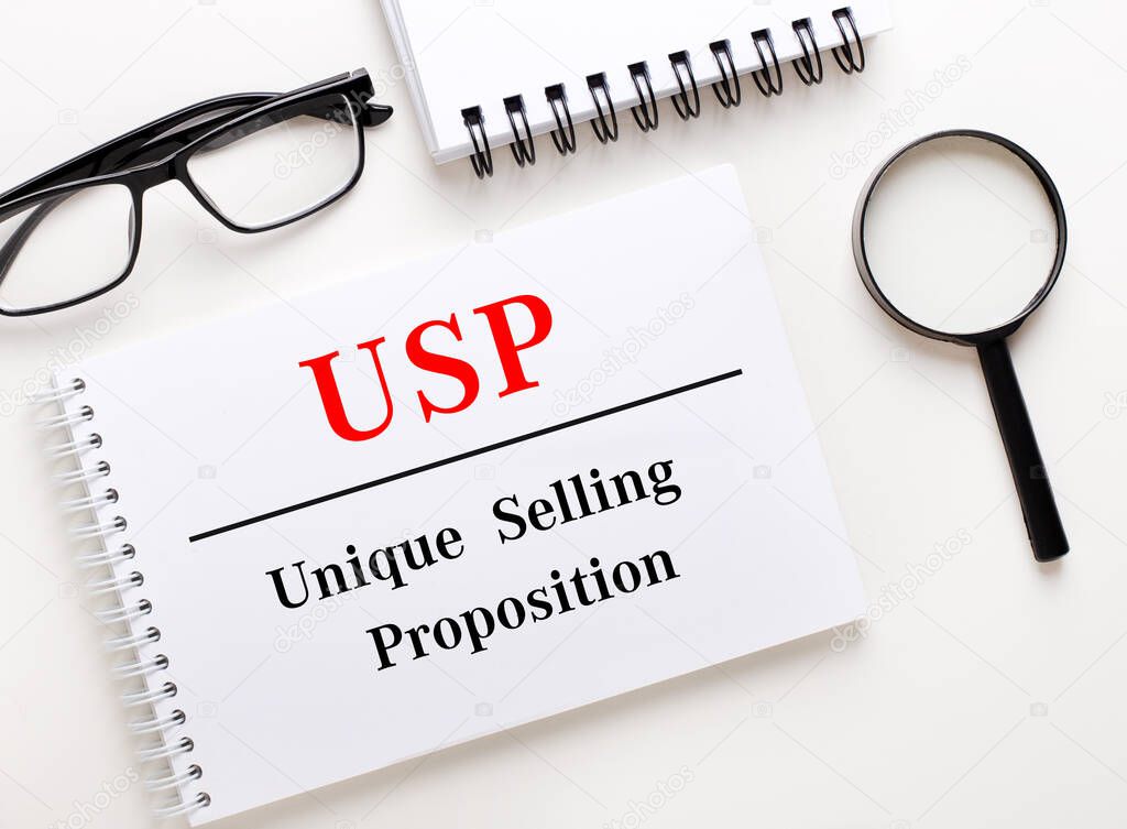 USP Unique Selling Proposition is written in a white notebook on a light background near the notebook, black-framed glasses and a magnifying glass.