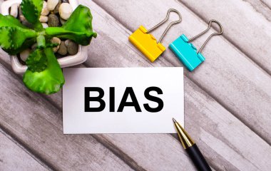 Tahta bir arka planda, BIAS yazılı beyaz bir kart, sarı ve yeşil ataçlar ve saksıda bir bitki. Yukarıdan görüntüle