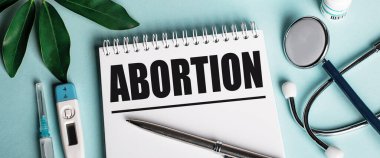 Mavi arka planda beyaz bir defterde, bir katman kılıfın yanında, bir steteskop, bir şırınga ve bir elektronik termometre ile, ABORTION kelimesi yazılır. Tıbbi konsept