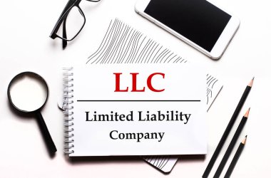 Beyaz bir arka plan gözlüğü, bir büyüteç, kalemler, bir akıllı telefon ve LLC Limited Limibility Company yazılı bir defter.