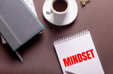Kahverengi arka planda, günlükler, bir fincan beyaz kahve ve MindSET 'li bir defter.