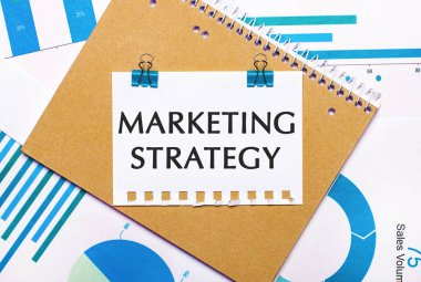 Masaüstünde mavi ve açık mavi grafikler ve diyagramlar, kahverengi bir defter ve mavi raptiyeli bir sayfa ve MARKETING STRATEGY metni bulunmaktadır. Yukarıdan bak. İş konsepti