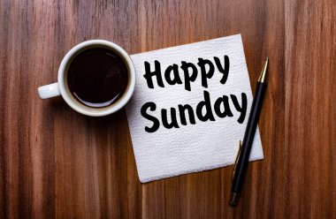 On a wooden table next to a white cup of coffee and a pen is a white paper napkin with the words HAPPY SUNDAY