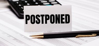 Masanın üzerinde raporlar, bir kalem, bir hesap makinesi ve POSTPONED yazılı bir beyaz kart var. Odaklanmayın