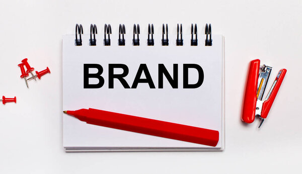 На светлом фоне красная ручка, красный степлер, красные скрепки и блокнот с надписью Бренд