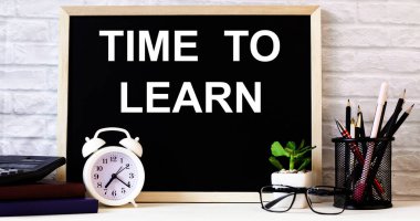 Time to Learn kelimeleri beyaz alarm saati, gözlük, saksı bitkisi ve kalem standının yanındaki tahtada yazılıdır..