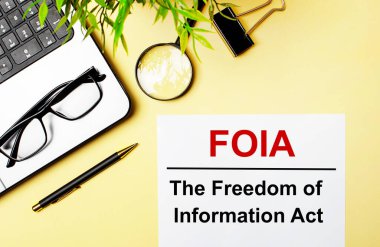 FOIA Bilgi Özgürlüğü Yasası kırmızı bir kağıt parçasına açık sarı bir arka planda dizüstü bilgisayar, kalem, büyüteç, gözlük ve yeşil bir bitkinin yanında yazılır..