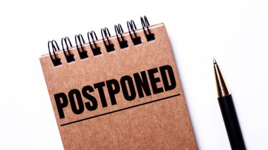 Açık bir arkaplanda, siyah bir kalem ve siyah yaylar üzerine kahverengi bir defter. Üzerinde POSTPONED yazılıydı.