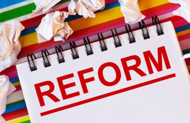 Çok renkli bir arkaplanda beyaz kağıt parçaları ve üzerinde kırmızı REform yazılı beyaz bir defter var.