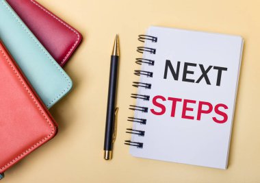 Çok renkli günlükler bej bir arka planda bir kalem ve NEXT STEPS harflerinin olduğu bir defterin yanında yatar. Düz yatıyordu.