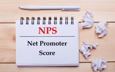 Tahta bir arka planda, NPS Net Promoter Puanı yazılı beyaz bir defter, beyaz bir kalem ve buruşuk beyaz kağıt parçaları