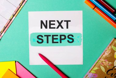 Parlak çıkartmalar, not defterleri ve işaretlerin yanında turkuaz bir arkaplan üzerine yazılmış NEXT STEPS