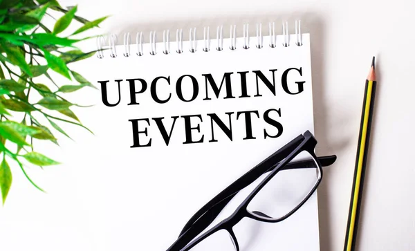 UPCOMING EVENTS is written in a white notebook next to a pencil, black-framed glasses and a green plant.