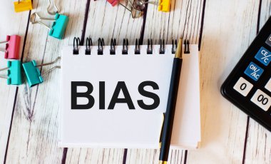 Açık ahşap bir masada hesap makinesi, çok renkli ataçlar ve kalemli bir defter ve BIAS sözcüğü bulunur.