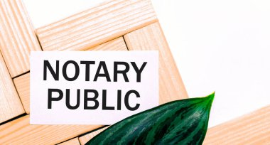 Beyaz arkaplanda tahta yapı taşları, üzerinde NOTARY PUBLIC yazılı beyaz bir kart ve bitkinin yeşil bir yaprağı. Yukarıdan görüntüle