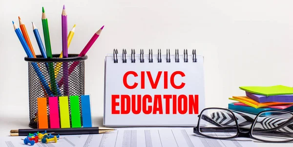 Masanın üzerinde renkli kalemler, parlak renkli çıkartmalar, gözlükler ve CIVIC EDUCATION yazılı bir defter var.