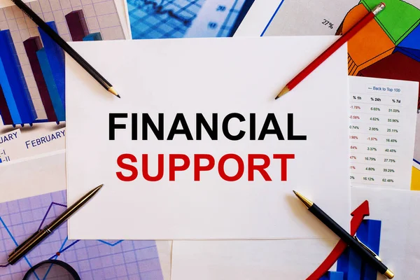 The words FINANCIAL SUPPORT is written on a white background near colored graphs, pens and pencils. Business concept