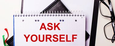 The words ASK YOURSELF is written in red in a white notebook next to black-framed glasses.
