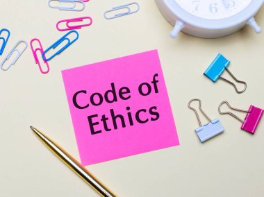 Hafif bir arka planda, beyaz bir çalar saat, pembe, mavi ve beyaz ataçlar, altın bir kalem ve ETHICS 'in metin kodlu pembe bir çıkartma.