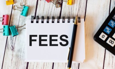 Hafif bir ahşap masada bir hesap makinesi, çok renkli ataçlar ve kalemle birlikte bir defter bulunur.