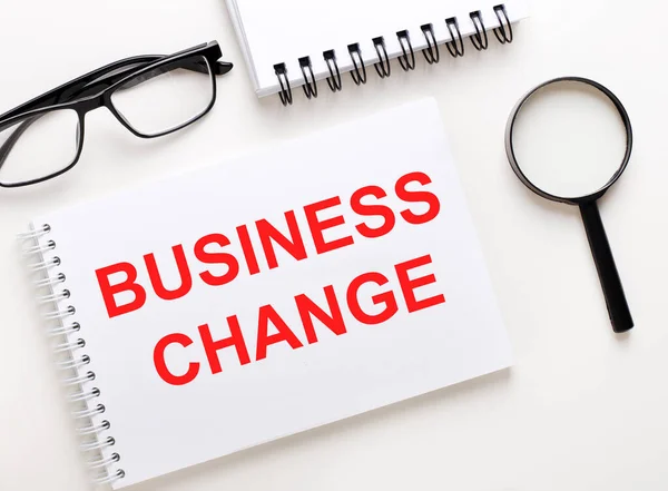 BUSINESS CHANGE is written in a white notebook on a light background near the notebook, black-framed glasses and a magnifying glass.