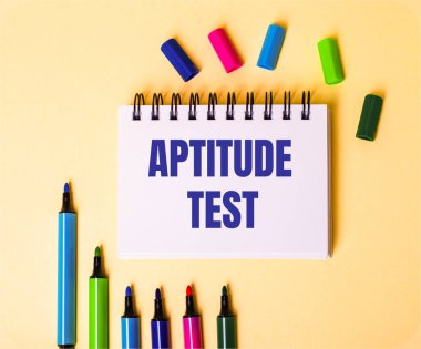 APTITUDE TEST kelimeleri çok renkli işaretlerin yanında bej bir arkaplan üzerine beyaz bir not defterine yazıldı