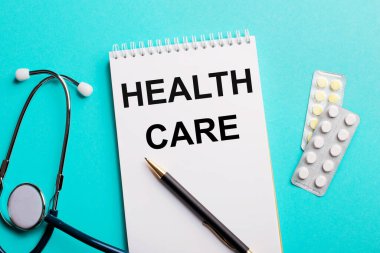 Stetoskop yakınında beyaz bir not defterine yazılmış sağlık bakımı, açık mavi arka planda kalemler ve haplar. Tıbbi konsept