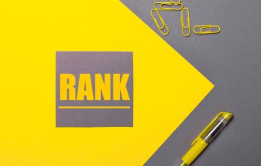 Gri ve sarı arka planda sarı renkli gri bir çıkartma RANK, sarı ataç ve sarı bir kalem var.
