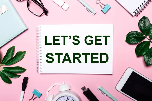 Let 's Get Başladı beyaz bir deftere yazılmıştır pembe arka plan üzerinde iş aksesuarları ve yeşil yapraklar ile çevrili.