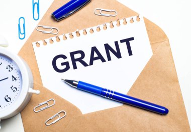 Hafif bir arka planda, bir Kraft zarfı, bir çalar saat, ataçlar, mavi bir kalem ve içinde GRANT olan bir sayfa kağıt..