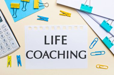 Hafif bir arka planda, bir yığın belge, bir beyaz hesap makinesi, sarı ve mavi ataçlar ve mandallar ve LIFE COACHING metin bir defter vardır