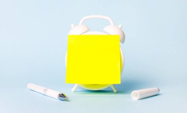 Açık mavi bir arka planda beyaz bir kalem ve üzerine yazı yapıştırılabilecek parlak sarı bir çıkartma yapıştırılmış bir çalar saat var. Şablon. Boşluğu kopyala.