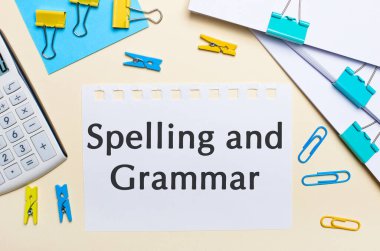 Hafif bir arkaplanda, bir yığın belge, bir beyaz hesap makinesi, sarı ve mavi ataçlar ve mandallar ve içinde Heceleme ve GRAMMAR yazılı bir defter vardır.