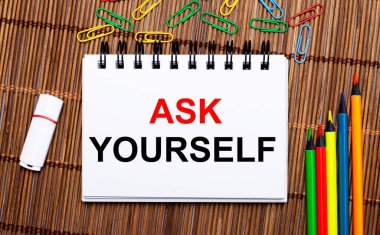 Tahta bir masada, çok renkli kalemler, ataçlar, beyaz bir flaş bellek ve ASK YOURSELF yazılı bir defter. Düz yatıyordu