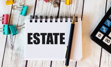 Açık ahşap bir masada hesap makinesi, çok renkli ataçlar ve kalemli bir defter bulunur.