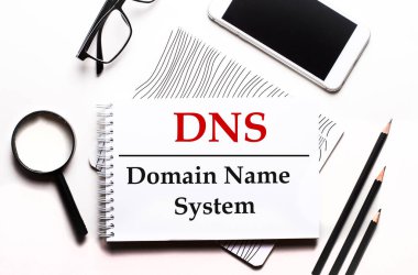 Beyaz bir arka plan gözlüğü, bir büyüteç, kalemler, bir akıllı telefon ve DNS Alan Adı Sistemi yazılı bir defter