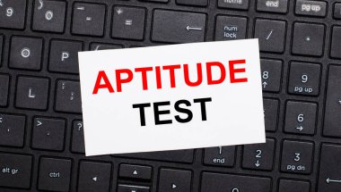 Siyah bir bilgisayar klavyesinde üzerinde APTITUDE TEST yazan beyaz bir kart var. Yukarıdan görüntüle
