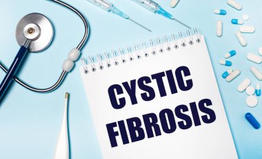 Açık gri arka plan, stetoskop, elektronik termometre, haplar, şırıngalar ve CYSTIC FIBROSIS yazılı bir defter. Tıbbi konsept.