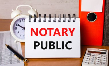 Masada raporlar, bir beyaz alarm saati, bir hesap makinesi, kağıtlar için klasörler, bir kalem ve üzerinde NOTARY PUBLIC yazan beyaz bir defter var. İş konsepti