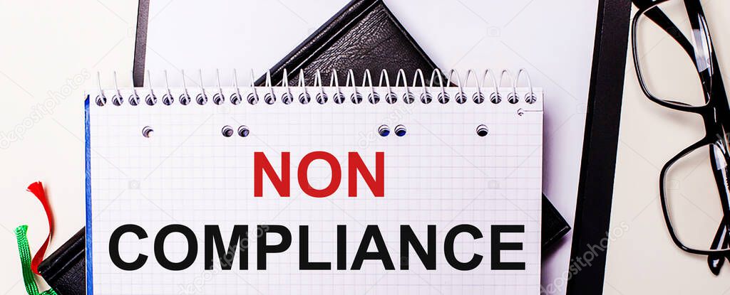 The words NON COMPLIANCE is written in red in a white notebook next to black-framed glasses.