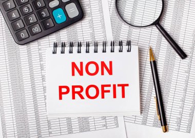 Masanın üzerinde raporlar, bir büyüteç, bir hesap makinesi, bir kalem ve NON PROFIT yazılı beyaz bir defter var. İş konsepti
