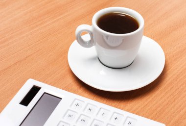 Tahta bir masanın üzerinde beyaz bir fincan kahve ve beyaz bir hesap makinesi var. İş konsepti