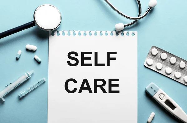 The words SELF CARE written on a white notepad on a blue background near a stethoscope, syringe, electronic thermometer and pills. Medical concept