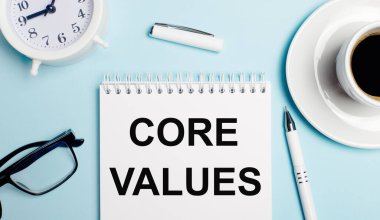 Açık mavi arka planda, kahveli beyaz bir fincan, beyaz bir çalar saat, beyaz bir kalem ve CORE VALUES yazılı bir defter. Yukarıdan görüntüle