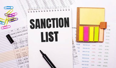 SANCTION LIST kelimelerini içeren bir defter, bir işaret, renkli ataç ve parlak not kağıdı grafiklerin arka planında yatıyor.