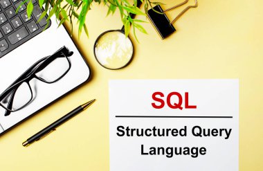 SQL Yapılandırılmış Sorgu Dili, bir dizüstü bilgisayar, kalem, büyüteç, gözlük ve yeşil bitkinin yanında açık sarı arkaplan üzerinde beyaz bir kağıt parçasına kırmızı ile yazılmıştır..